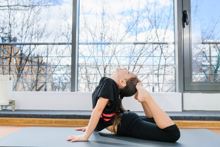 Preteen flexible girl lay on tummy and touch back of head by feet at light room with panoramic window, gymnastics for kidsの写真素材