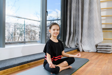 Preteen gymnast smiling child girl sit on sport mat in lotus yoga pose near panoramic window, healthy childhoodの写真素材
