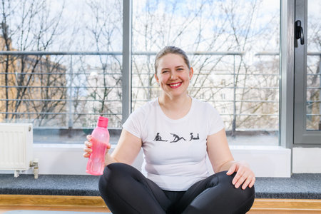 Body positive woman sit in yoga lotus pose with sport water bottle and smile in light fitness studioの写真素材