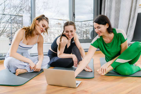 Three sportive girls sit on mats in fitness studio during online yoga exercises workout training tutorialsの写真素材