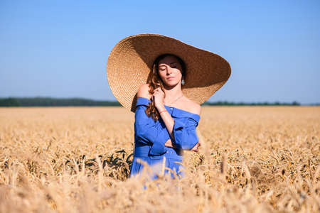 Adorable young woman stay in retro blue dress and wide brimmed hat inside wheat field, countryside romantic fashionの写真素材