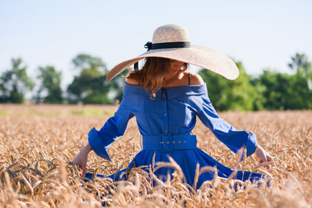 Romantic fashion girl stand in wheat field, wearing blue vintage dress with belt and wide brim straw hatの写真素材