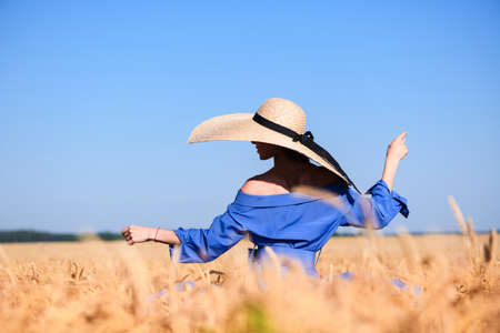 Girl stay back in wide brim hat and blue dress inside summer wheat field, coutryside vintage fashion. Woman dancing among the ears.の写真素材