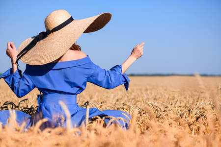Girl stay back in wide brim hat and blue dress inside summer wheat field, coutryside vintage fashion. Back view.の写真素材