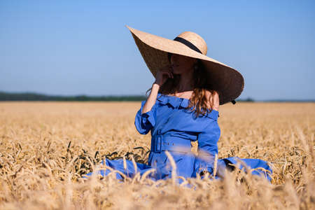 Romantic fashion girl stand in wheat field, wearing blue vintage dress with belt and wide brim straw hatの写真素材