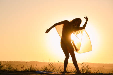 Beautiful girl silhouette dancing exercise against warm sun in transparent cloth, female fit body curvesの写真素材
