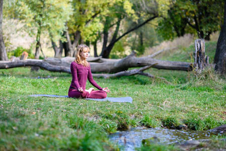 Woman sit on sport mat in lotus yoga meditation pose at beautiful green forest and water stream nature backgroundの写真素材