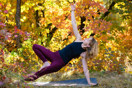 Woman middle age doing yoga balance training workout in side plank pose on mat at autumn forest, healthy lifestyleの写真素材