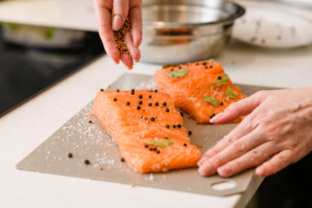 Female hand sprinkles a piece of salmon fillet lying on a cutting board with spicesの写真素材