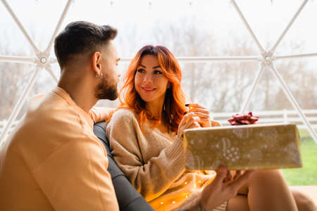 Young couple are sitting in a cozy glamping. The man gives a gift to the woman for birthday. Huge window in the backgroundの写真素材