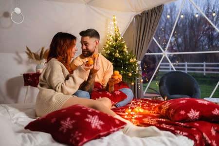 Cheerful young couple are playing with tangerines while sitting on the bed on Christmas evening.の写真素材