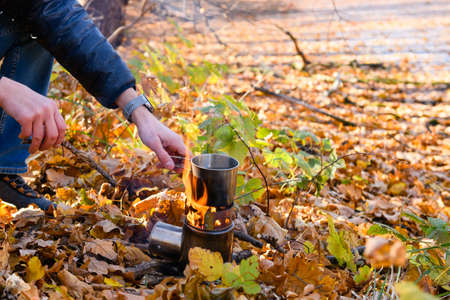 A tourist heats water in a metal mug on a fire in an autumn forest. Hand holding a mug on fire.の写真素材