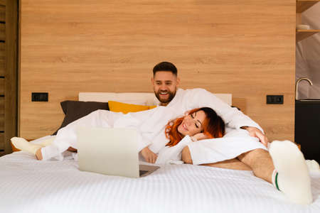 A young couple in bathrobes is lying on the bed with a laptop. Man and woman watching a movie. A woman lies with her head in the lap of a man.の写真素材