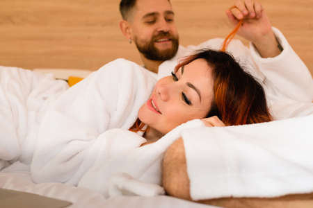 Close-up of beautiful young couple in bathrobes lying together on bed. A man playing with a woman's hair. A woman lies with her head in the lap of a man.の写真素材