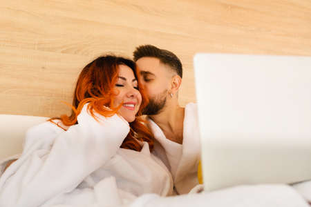 Happy young couple in bathrobes lie in bed with laptop. A man kissing a woman.の写真素材