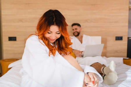Portrait of a smiling attractive young woman with red hair in a white bathrobe sitting on the bed.の写真素材
