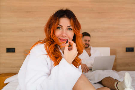 Portrait of a smiling attractive young woman with red hair in a white bathrobe sitting on the bed. The girl looking at the camera.の写真素材
