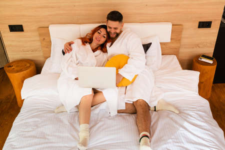Full length view of a young couple in bathrobes lying on the bed with a laptop. Man and woman watching a movie. View from above.の写真素材
