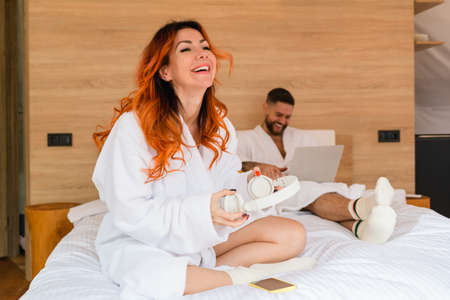 Woman with headphones laughs sitting cross-legged on the bed. Red-haired woman in a bathrobe with headphones.の写真素材