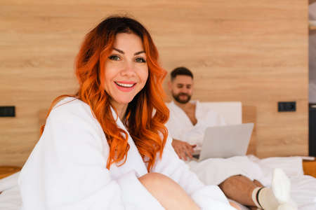 Portrait of a smiling attractive young woman with red hair in a white bathrobe sitting on the bed. Close-up.の写真素材