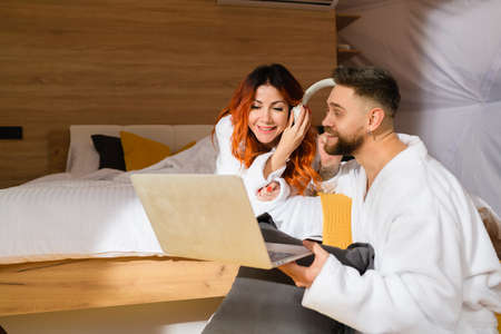 Photo of a young couple in bathrobes lying on the bed with a laptop. Man and woman listen to music.の写真素材