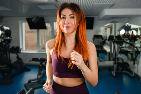 Positive woman exercise in gym treadmill, red-eared female running at track at sports hall interiorの写真素材