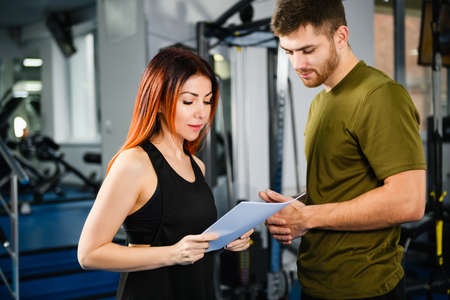 Fit woman customer client reading paper gym documents, instructions and making contract with handsome trainer coachの写真素材