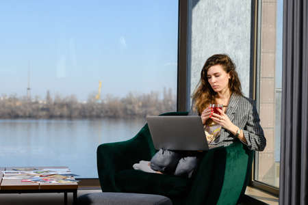 Business woman sit with laptop and cup of tea in cozy home interior, modern apartment with panoramic window and river viewの写真素材