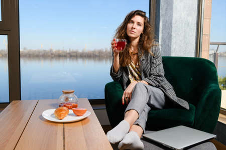 Woman in cozy home atmosphere, breakfast before remote job, sitting in armchair near panoramic window and table with fruit tea and croissant, hold cup and laptopの写真素材