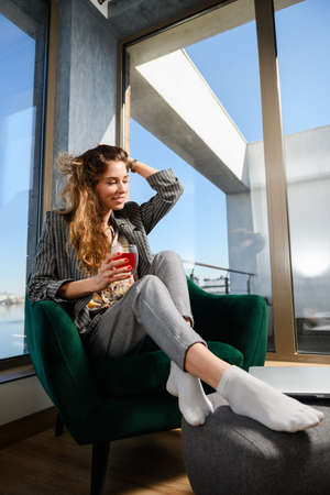 Girl freelancer in modern flat of glass and concrete sitting at cozy armchair with laptop, hold cup of tea, remote job and luxury livingの写真素材