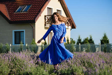 Attractive young positive woman in a blue dress is spinning, dancing in a lavender field. country house in the background. Hair and dress are blowing in the wind.の写真素材