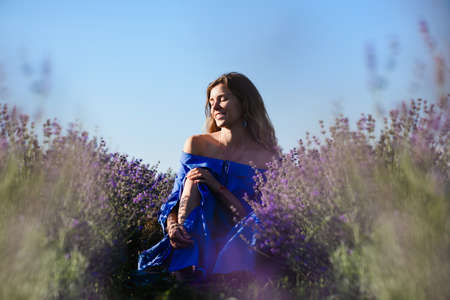 An attractive young woman with bare shoulders sits between rows of lavender. Girl smiling with closed eyes. Blue sky in the background.の写真素材