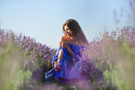 An attractive young woman with bare shoulders sits between rows of lavender. Girl hugging herself sensually. Blue sky in the background.の写真素材