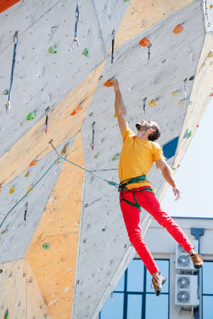 Sportsman climber hangs on one hand on an artificial climbing wall outdoors. Young and fit man exercise free mountain climbing on outdoor practice wall.の写真素材