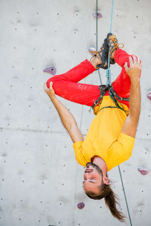 A man hangs upside down in a lotus position on a climbing wall. The man is wearing a yellow T-shirt...