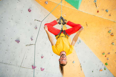 A man hangs upside down in a lotus position on a climbing wall. The man is wearing a yellow T-shirt and red pants.の写真素材