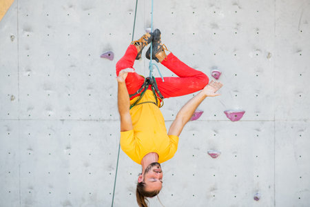A man hangs upside down in a lotus position on a climbing wall. The man is wearing a yellow T-shirt and red pants.の写真素材