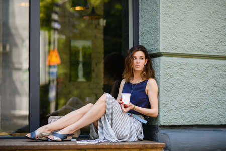 Young attractive woman enjoying takeaway coffee while sitting by a huge window outdoors. The girl is holding a disposable cup.の写真素材