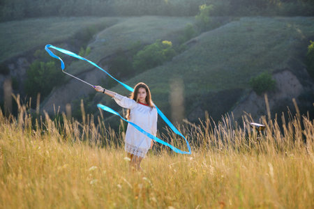 A teenage girl with a gymnastic ribbon on a hillside. girl on the background of bayrak. Tall dry wild grass.の写真素材