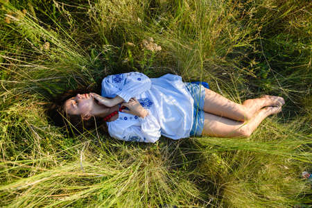A teenage girl lies in a tall meadow grass. The girl is dressed in a national Ukrainian shirt -...