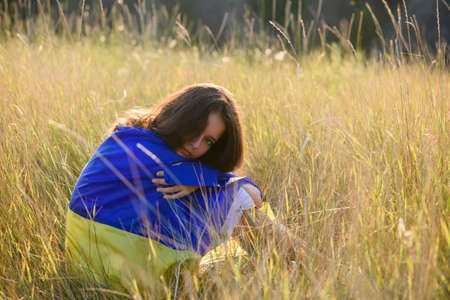 A teenage girl sits in tall wild grass on the slope of a ravine with a Ukrainian flag on her shoulders.の写真素材
