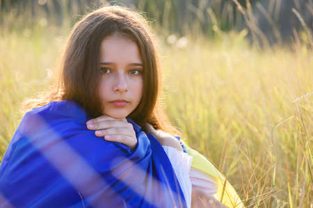 A teenage girl sits in tall wild grass on the slope of a ravine with a Ukrainian flag on her shoulders.の写真素材