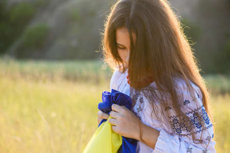 A sad teenage girl holds a Ukrainian flag to her chest. The girl is dressed in Ukrainian national clothes - vyshyvanka. Soft backlight at sunset.の写真素材