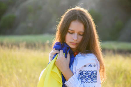 A sad teenage girl holds a Ukrainian flag to her chest. The girl is dressed in Ukrainian national clothes - vyshyvanka. Soft backlight at sunset.の写真素材