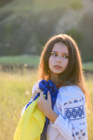 A sad teenage girl holds a Ukrainian flag to her chest. The girl is dressed in Ukrainian national clothes - vyshyvanka. Soft backlight at sunset.の写真素材
