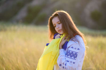 A sad teenage girl holds a Ukrainian flag to her chest. The girl is dressed in Ukrainian national clothes - vyshyvanka. Soft backlight at sunset.の写真素材