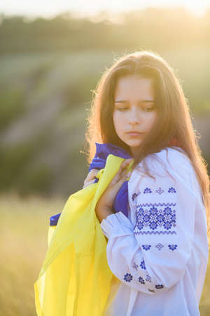 A sad teenage girl holds a Ukrainian flag to her chest. The girl is dressed in Ukrainian national...