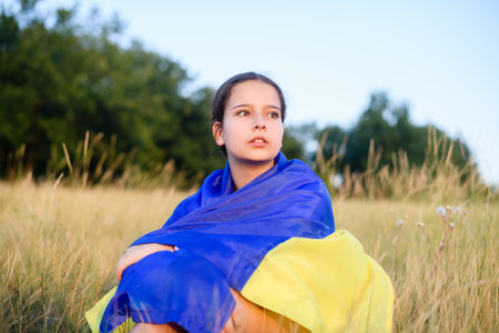A teenage girl sits in tall wild grass on the slope of a ravine with a Ukrainian flag on her shoulders.の写真素材
