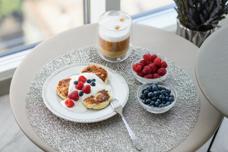 Cottage cheese pancakes for breakfast with blueberries and raspberries and sour cream. Cup of latte with delicious breakfast. Table with breakfast by the window on the background of the city.の写真素材