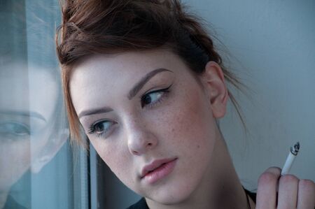 Beautiful young woman in casual wear smoking on window. Over evening  city.の写真素材